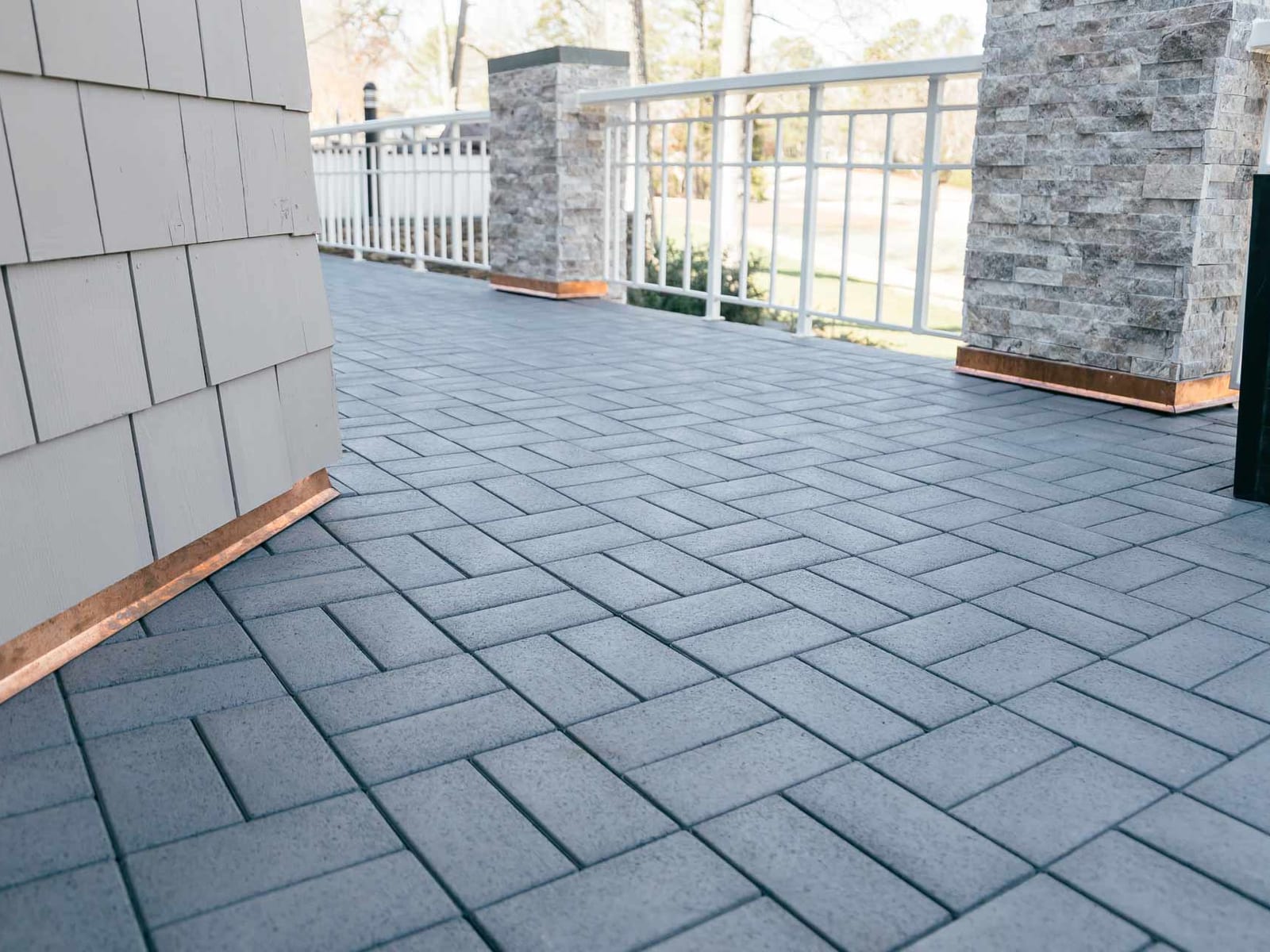Gray composite pavers on a covered balcony beside shake siding with copper trim at the base