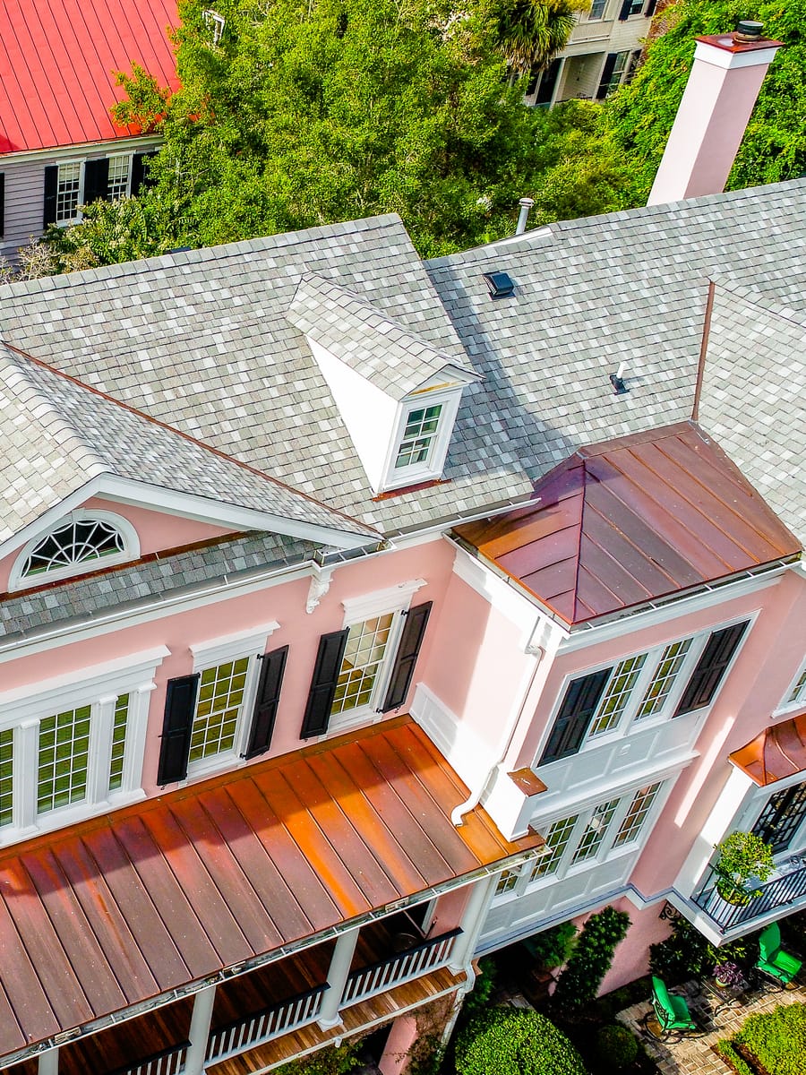 Slate roof with copper porch roofs on a pink Charleston home, beside red metal neighboring roofs